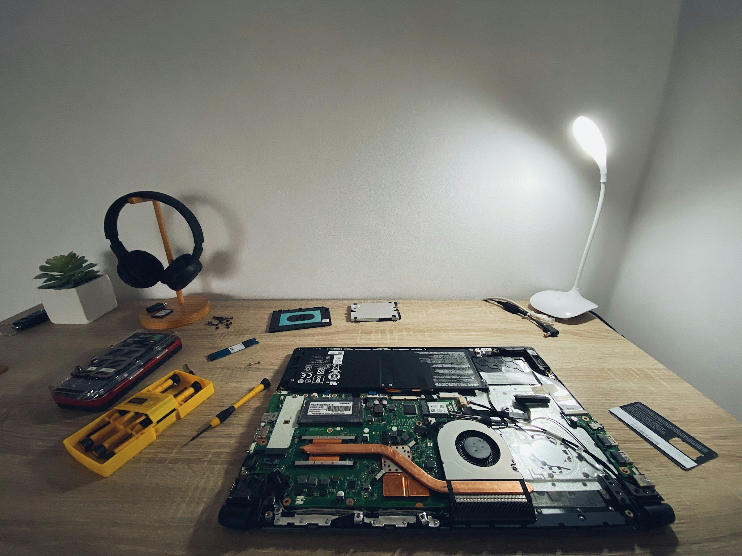 A repair bench with an opened laptop, screwdriver kit, spare drive and memory, and a desk lamp.